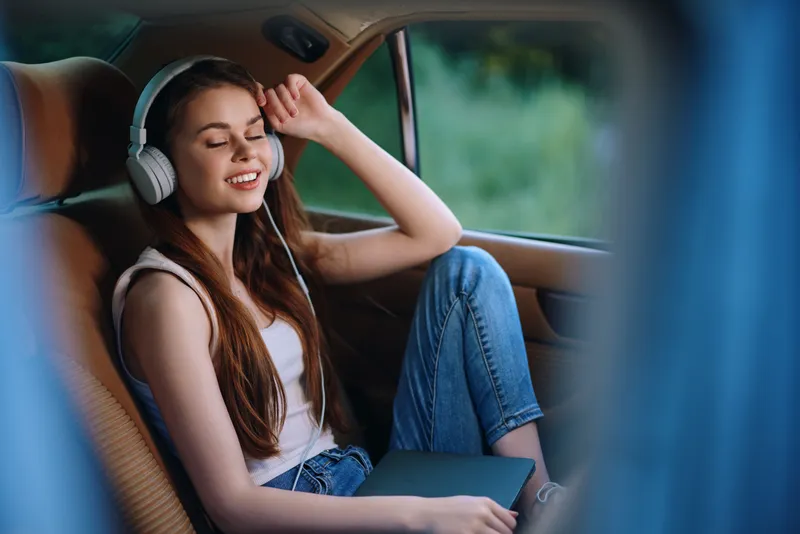 Young woman enjoying music in a car, wearing headphones, casual outfit, relaxed expression, green background, summer vibes, lifestyle concept.