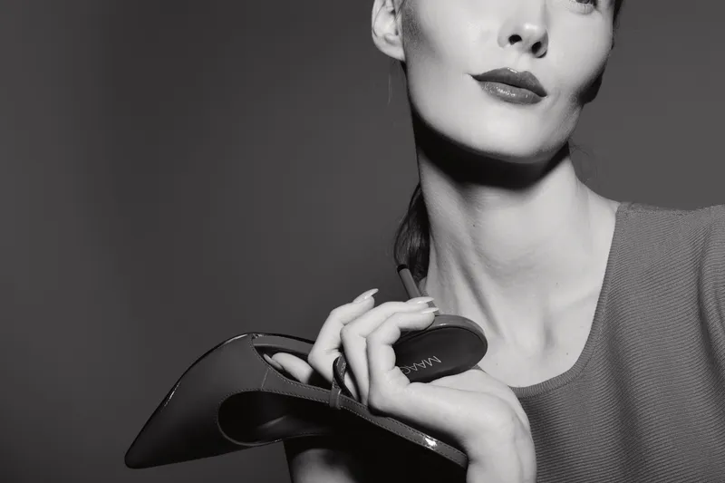 Closeup of a white woman with pale skin holding a black high heel shoe near her face in monochrome. Fashion and elegance concept with a vintage mood and soft lighting.
