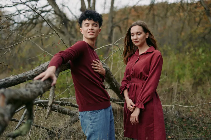 Two people stand outdoors beside a fallen tree, wearing burgundy clothing, expressing genuine connection and authenticity in a quiet, candid moment.