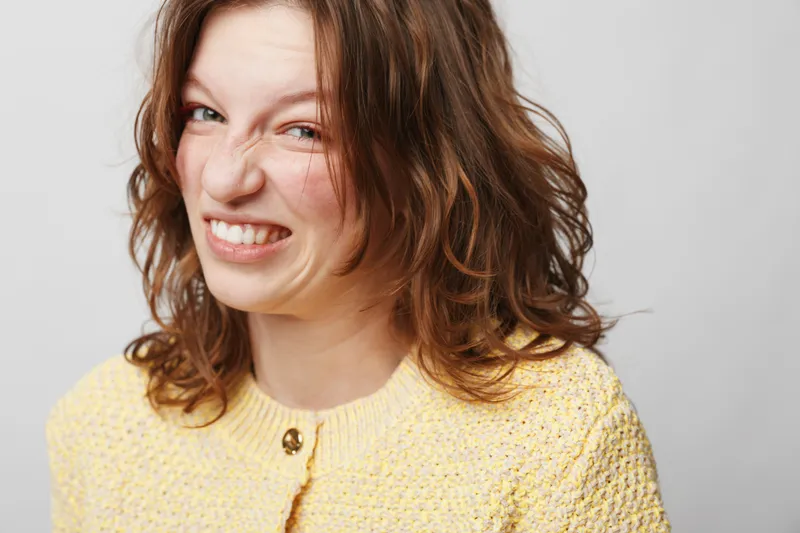 Young woman with curly brown hair wearing yellow sweater makes a funny grimace with scrunched face. Studio portrait on light background, concept of humor, emotion, and playful expression.