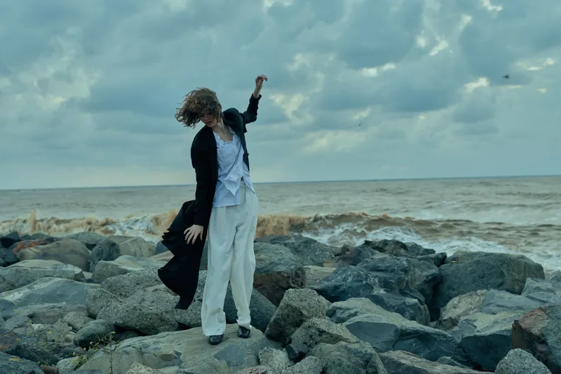 Woman on a rocky shore by the sea on a windy day, wearing a loose coat and white pants, exploring rugged coastline as waves crash and clouds roll.