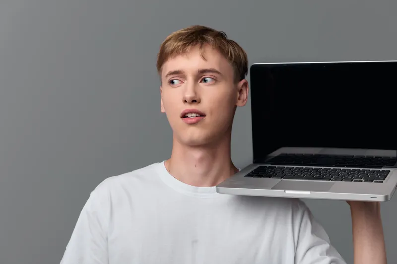 Casual young man with short hair holding a laptop beside his head, modern tech concept for remote work, minimal studio background, relaxed pose, digital device focus, clean composition and everyday