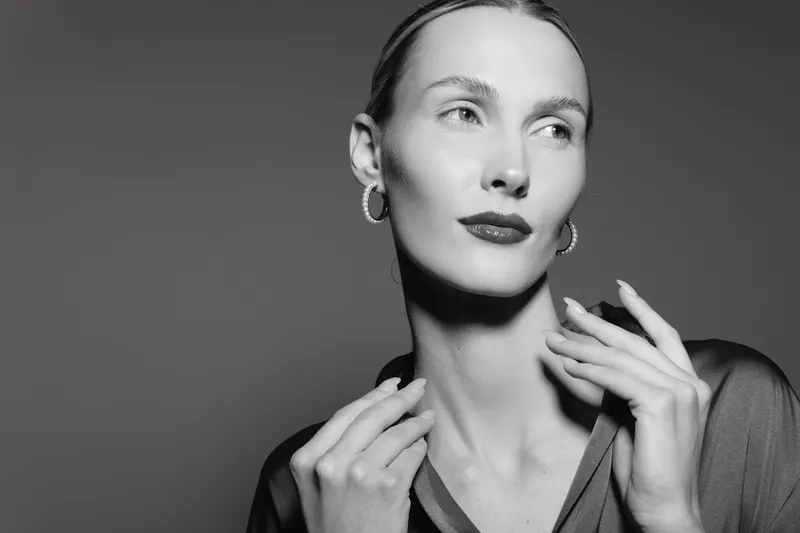 A slim white woman with pale skin and slicked hair poses elegantly looking sideways with hands near her neck in a monochrome studio. Concept of fashion, beauty, and portrait.