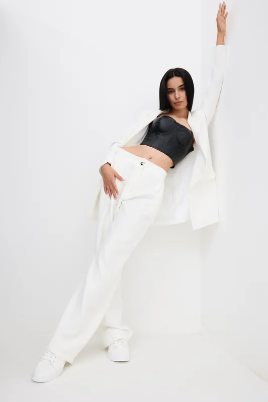 Young woman wearing white pants and blazer with black corset posing stylishly against a white background in a minimalist studio setting, showcasing modern fashion.