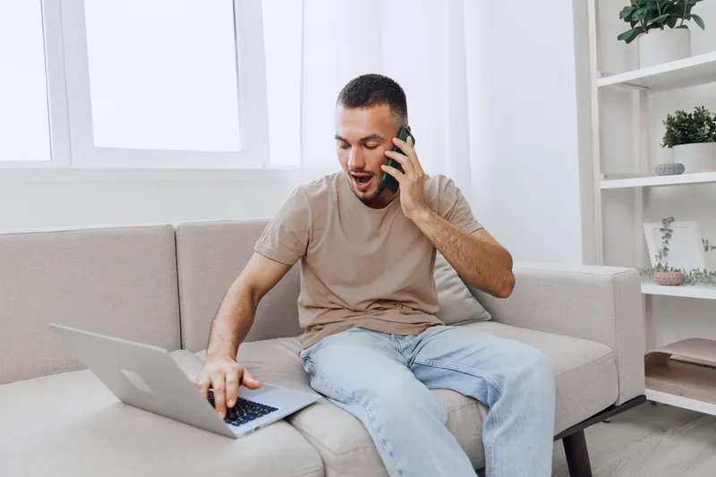 Young man engaged in a phone conversation while using a laptop, expressing excitement and focus, set in a modern, bright living room with a cozy ambiance.