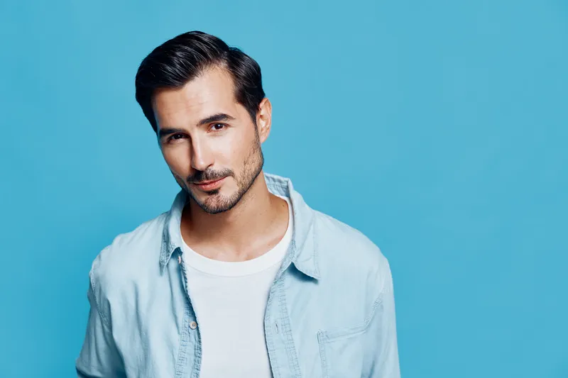Charming young man in a light denim shirt posing confidently against a vibrant blue background, offering a relaxed and approachable demeanor.