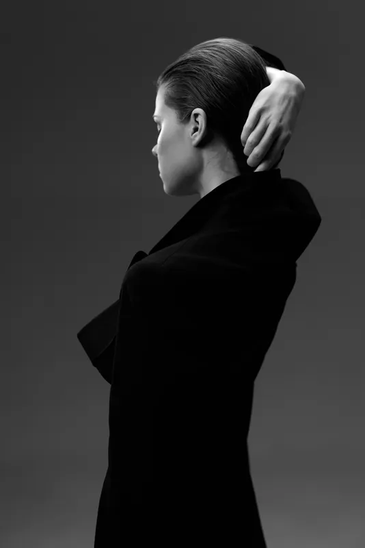 Black and white portrait of a woman with slicked-back hair wearing a dark coat and posing with her hand behind her head.