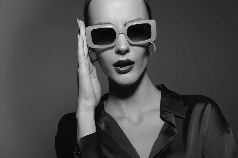 Elegant Caucasian woman with light skin wearing large square sunglasses and dark lipstick poses in studio with serious expression in black and white photo.