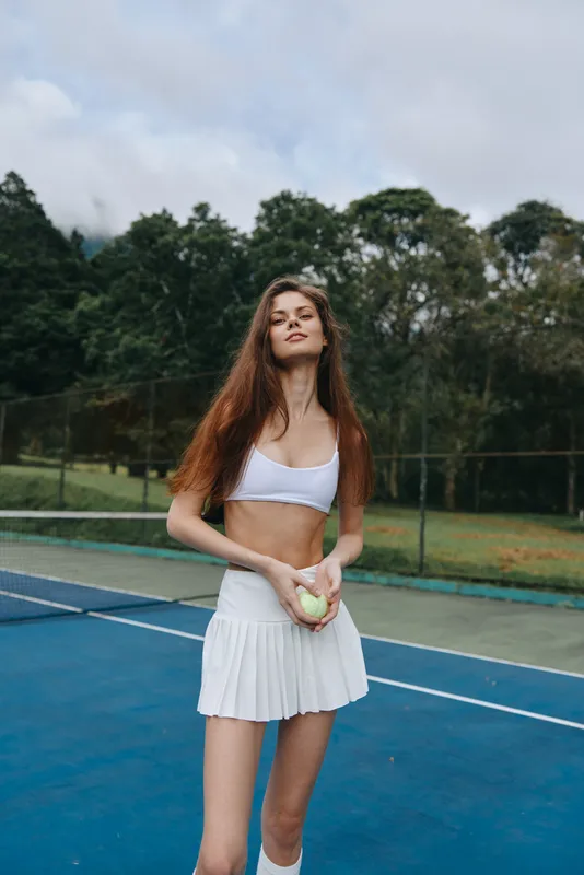 Athletic young woman holding a tennis ball on a sunny day, wearing a white crop top and a pleated white skirt, showcasing confidence and determination in a vibrant outdoor setting
