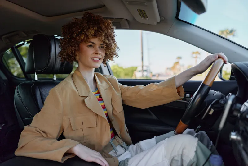 Smiling woman with curly hair driving a car, interior visible, warm light, everyday lifestyle moment, retro film color mood, evokes independence and self expression.