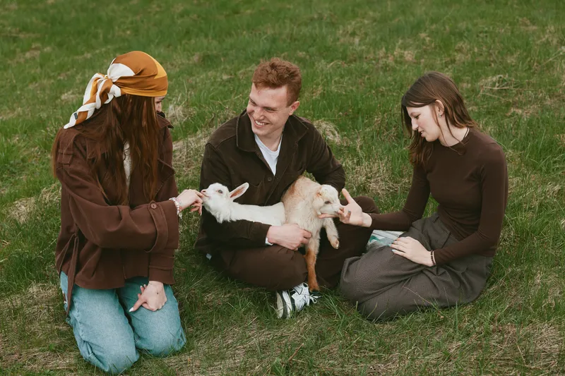 Cheerful group outdoors, adults and children share gentle contact with a goat kid, sunlit grass, casual clothing, relaxed smiles, natural colors, candid lifestyle moment, wholesome farming vibe