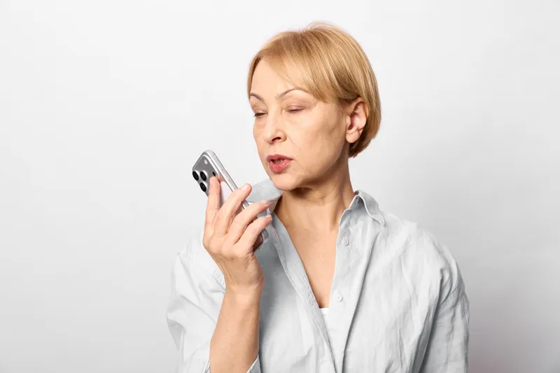 Woman with smartphone indoors, calling, communication, mature expression, studio portrait, white background, light blouse, modern technology, focused gaze, soft daylight, calm