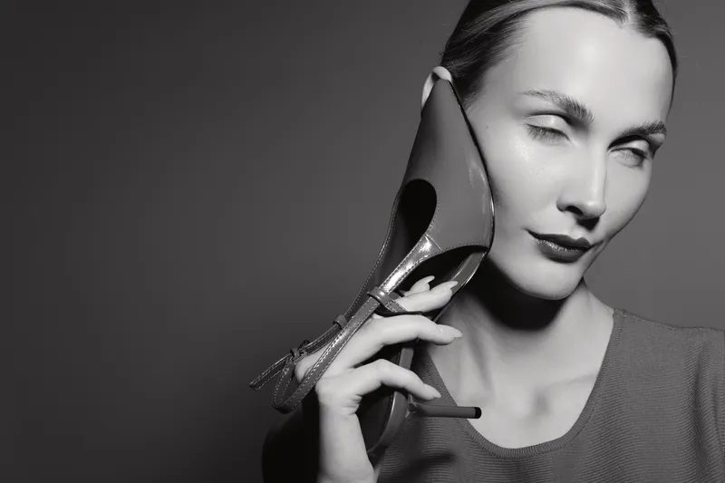 A fair skinned Caucasian woman holds a high heel shoe near her face in a black and white studio portrait.