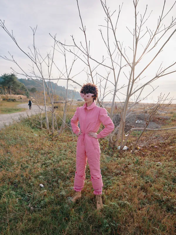Girl in pink jumpsuit standing in an outdoor landscape with trees, posing in a field, solitary figure amid natural scenery, confident and stylish in countryside setting