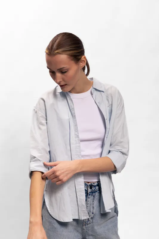 Young woman wearing casual shirt and white t-shirt looks down, touching her arm. Minimalist studio portrait on white background with natural expression and relaxed pose.
