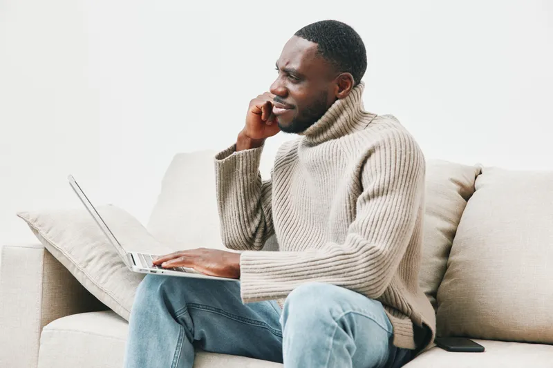 A concerned young African man in a cozy beige sweater talking on the phone while working on his laptop at home, expressing emotions of stress and focus