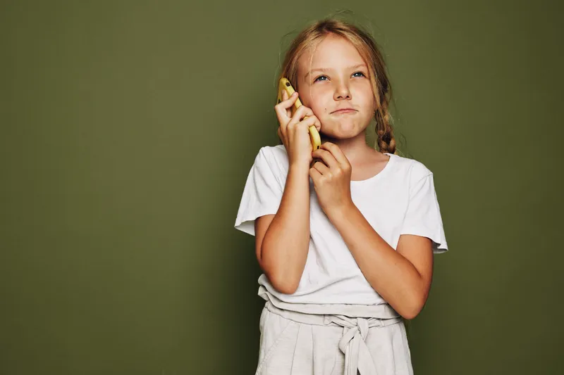 Thoughtful girl in a white t-shirt holds a banana to her ear as if talking. Olive background with soft lighting. Concept of imagination, childhood, and play.