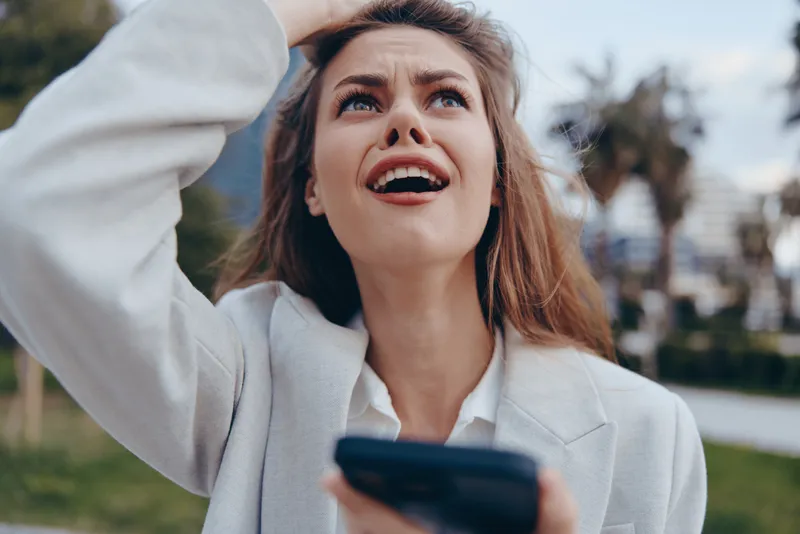 Businesswoman expressing frustration outdoors, wearing a light blazer and looking up with concern amidst a vibrant city backdrop with greenery.