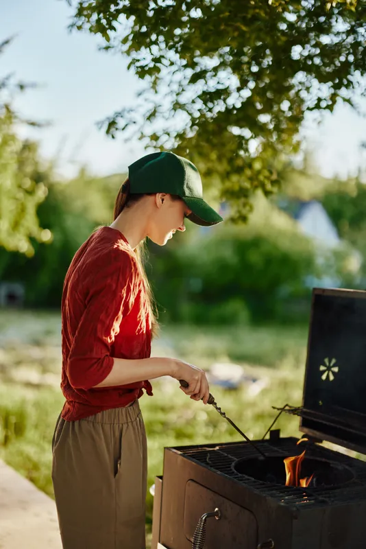 Young woman grilling outdoors in a green cap, wearing a red sweater and beige pants, evoking a joyful summertime experience amidst nature