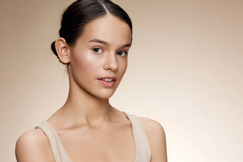 Portrait of a young woman with a natural look, showcasing delicate makeup and a neutral top against a soft beige background, ideal for beauty and skincare concepts