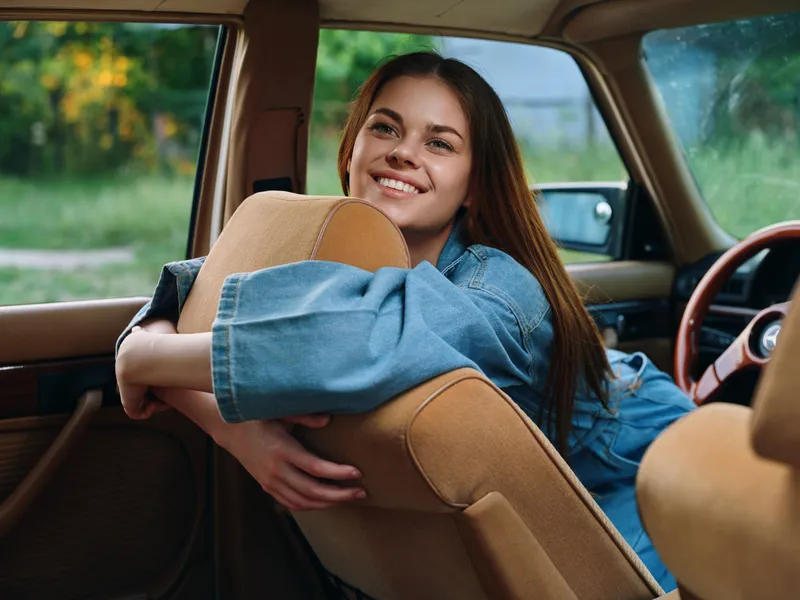 Cheerful young woman in a denim jacket relaxing in a vintage car, showcasing happiness and nostalgia in a green outdoor background.