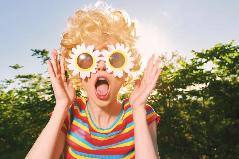 Joyful child wears daisy sunglasses and striped shirt, posing playfully outdoors under bright sun, capturing a sunny summer mood with colorful whimsical style.