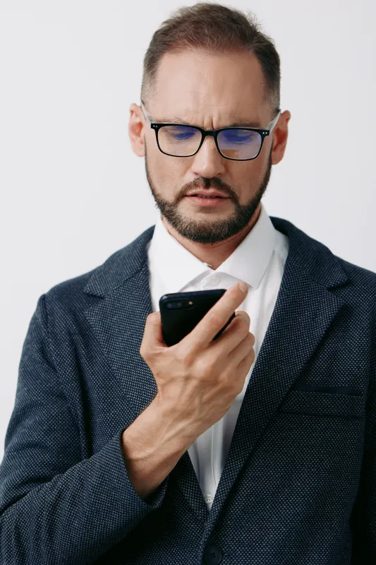 Professional businessman studies a mobile device, showing focus and determination. The portrait sits against a clean colored backdrop, isolating the subject and conveying confident mood.