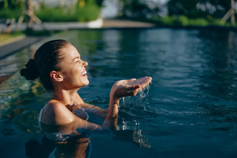 A woman in a pool enjoys a sunny moment, hands cupping water as she smiles. Bright outdoor light highlights a carefree, refreshing lifestyle moment by the water.