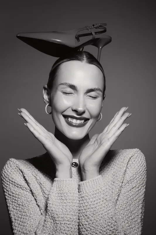 Smiling caucasian woman with light skin and earrings balances a high heel shoe on her head, posing with closed eyes and hands near her face in studio.