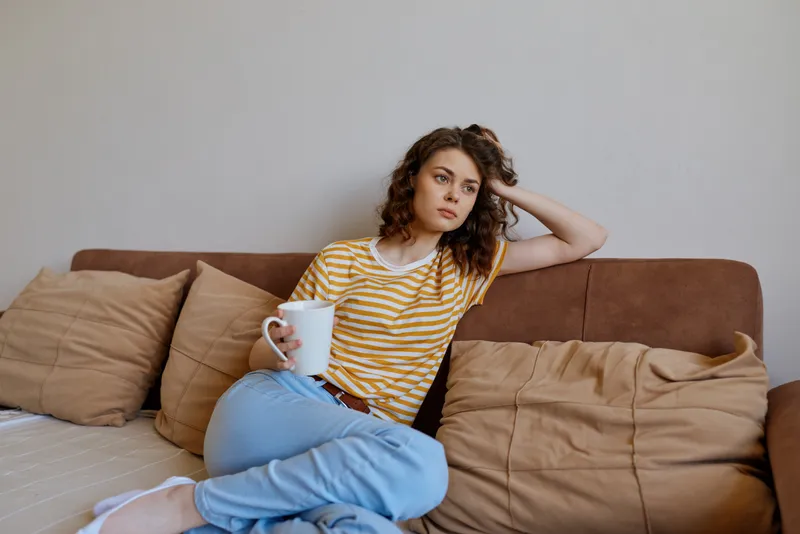 Casual woman relaxing at home on a couch with a mug, striped shirt and light jeans. Close up view of comfort, daily life, and self care in a cozy living space with soft lighting and serene mood.