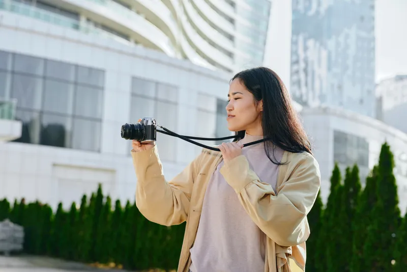 Woman with camera in an outdoor urban setting, capturing cityscape and modern architecture, casual jacket, calm expression, street photography mood, daylight framing, travel inspired, contemporary