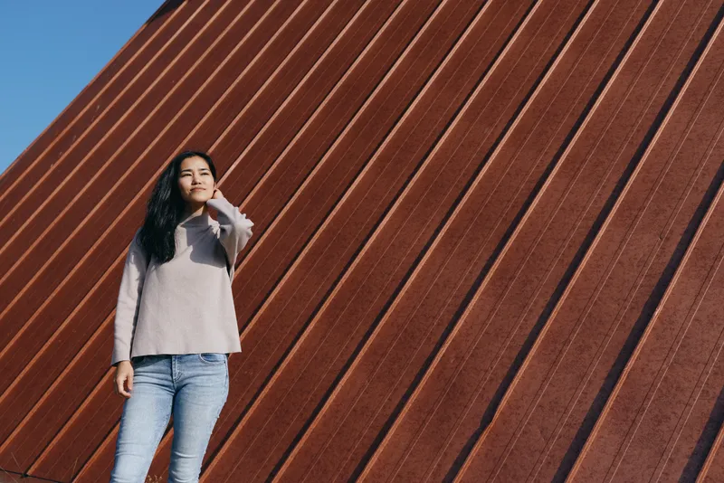 Woman standing against a tall diagonal wall in casual sweater and denim, bright outdoor light, urban backdrop, relaxed pose, street style photography with strong geometry and texture