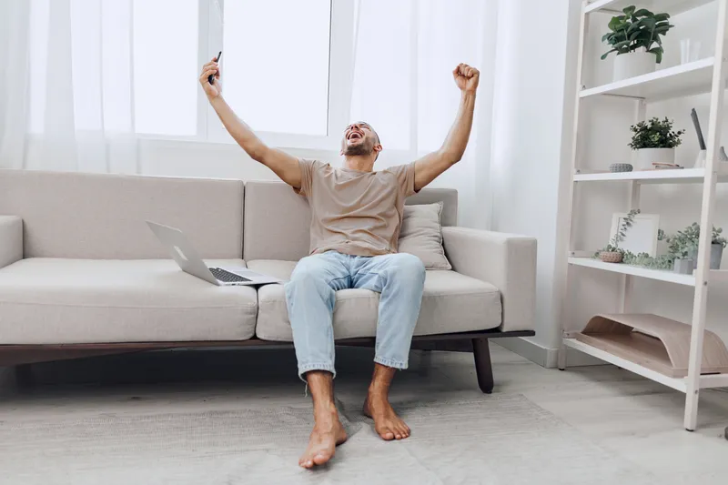 Excited man celebrating a successful moment at home, with a joyful expression while holding a smartphone. Emphasizing happiness and achievement in casual attire.