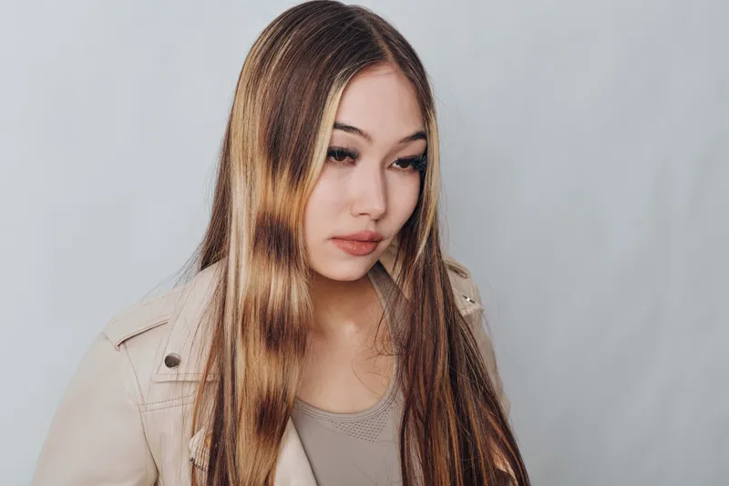Young woman with long hair and neutral makeup wearing beige jacket and grey top posing against light gray background studio portrait.
