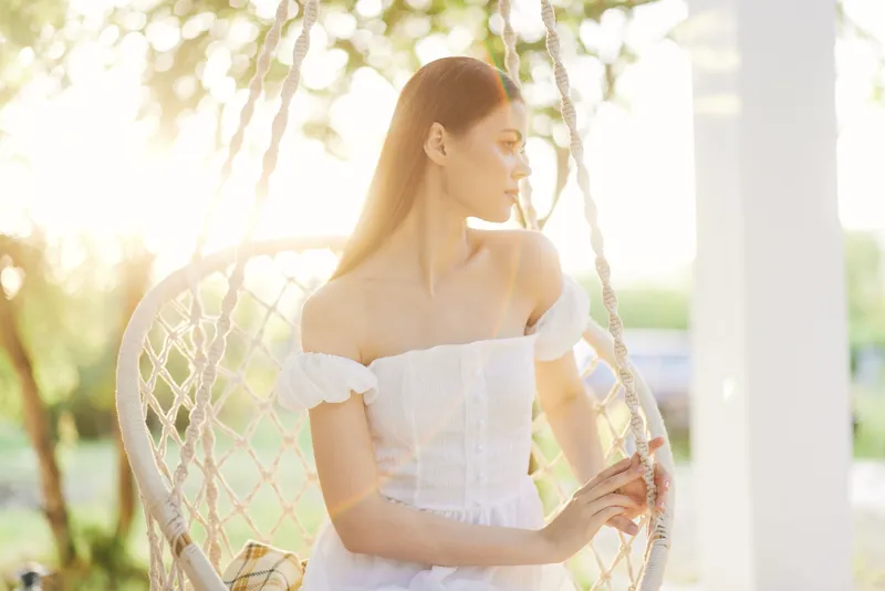 Young woman in white dress relaxing in a hanging chair, enjoying sunlight and nature, evoking feelings of serenity and peace during a warm season