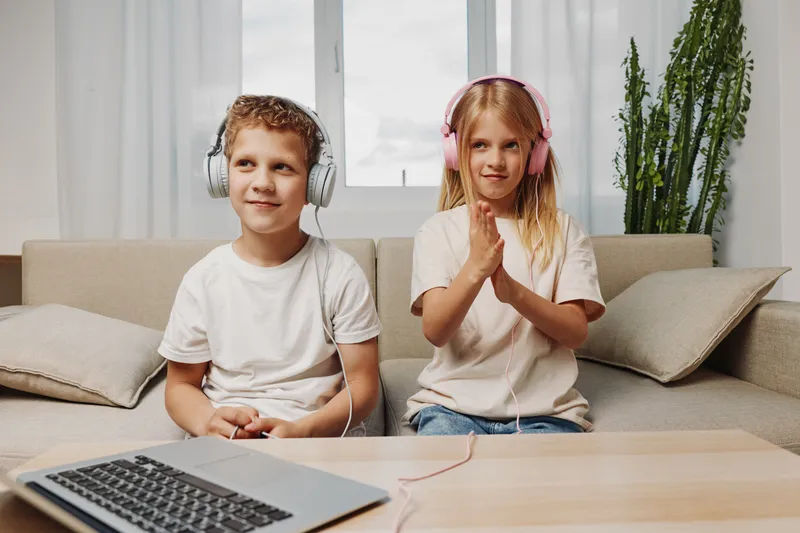 Two children wearing headphones sit on a beige sofa in a bright room with a window and green plant. The boy smiles looking away while the girl claps hands. Concept of music, childhood, leisure.