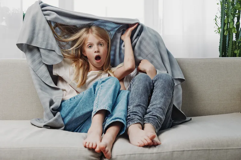 Two playful children sitting on a sofa covered with a blanket, one girl surprised with open mouth, casual home setting with natural light and relaxed mood.
