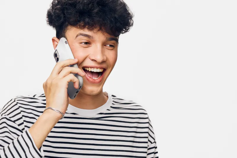 smiling man with curly hair talking on smartphone, wearing striped shirt, expressing happiness in bright studio setting with white background