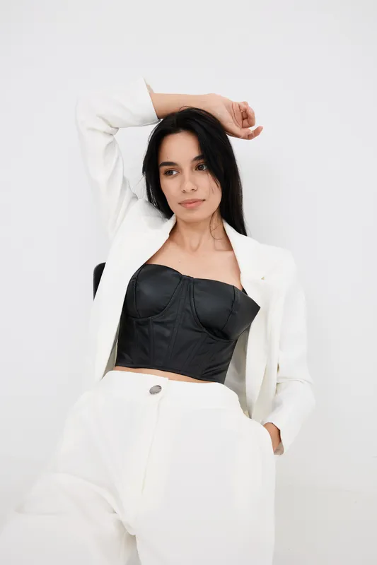 Young woman in stylish black corset and white suit posing casually against plain white background, showcasing fashionable modern outfit and relaxed confident mood.