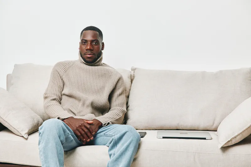 African man in a cozy turtleneck sweater sitting on a light colored sofa, reflecting calmness and introspection in a modern interior setting