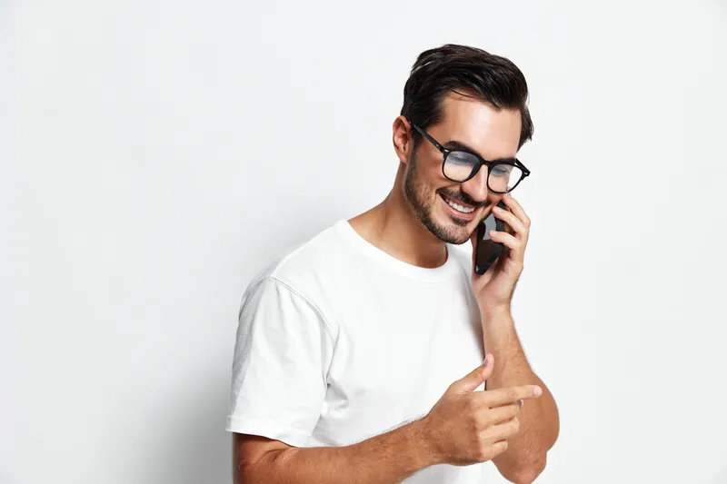 Happy smiling man talking on mobile phone while pointing at something, wearing casual white t-shirt, isolated on plain solid background for lifestyle portrait.