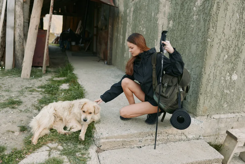 A cheerful traveler crouches to greet a friendly dog beside a rustic path, capturing a warm moment of connection and curiosity in a quiet outdoor setting that feels inviting and calm.