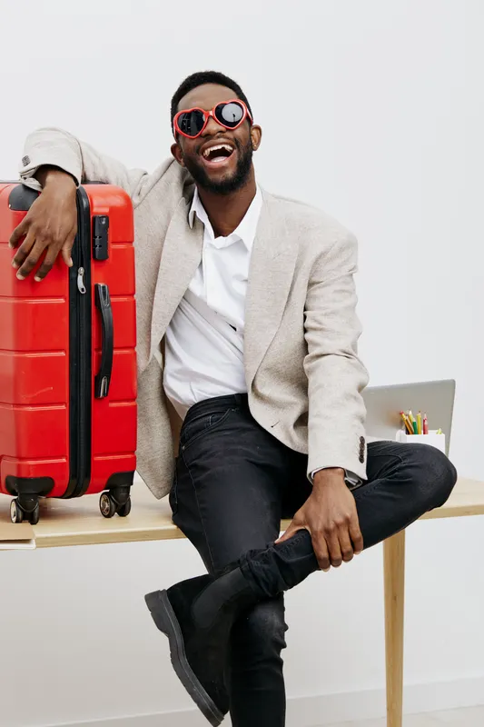 Smiling man with sunglasses holding red suitcase in casual clothing siting on table isolated on white background. Lifestyle, travel, happiness, vacation, fun, vacation concept