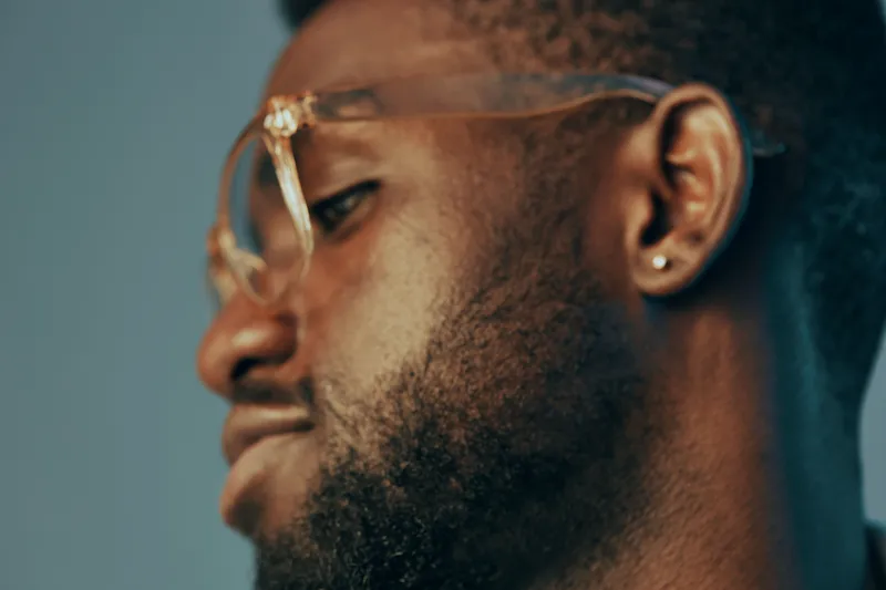 A close up portrait of a stylish Black man with glasses, showcasing a confident expression against a muted blue background, representing modern fashion and self assurance