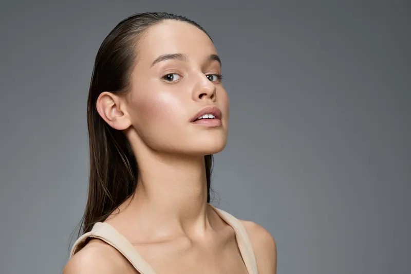 Natural beauty portrait of a young woman with long hair and radiant skin against a soft gray background, showcasing a fresh and minimalist style