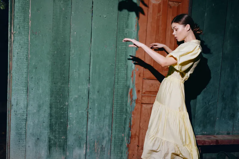 A woman in a pale yellow dress strikes a thoughtful pose beside a multicolored wall, soft lighting and shadow add an artistic mood to an outdoor setting.