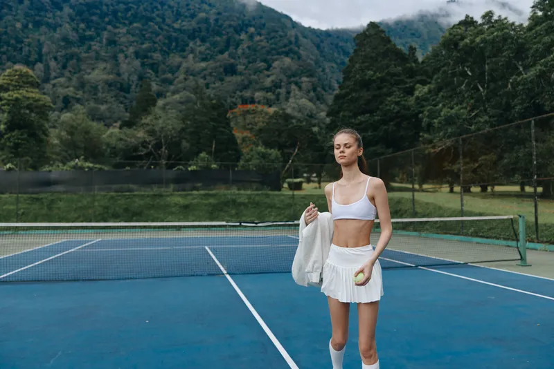 Young female tennis player in white athletic wear on a blue tennis court with a scenic mountain background, exuding confidence and determination after practice