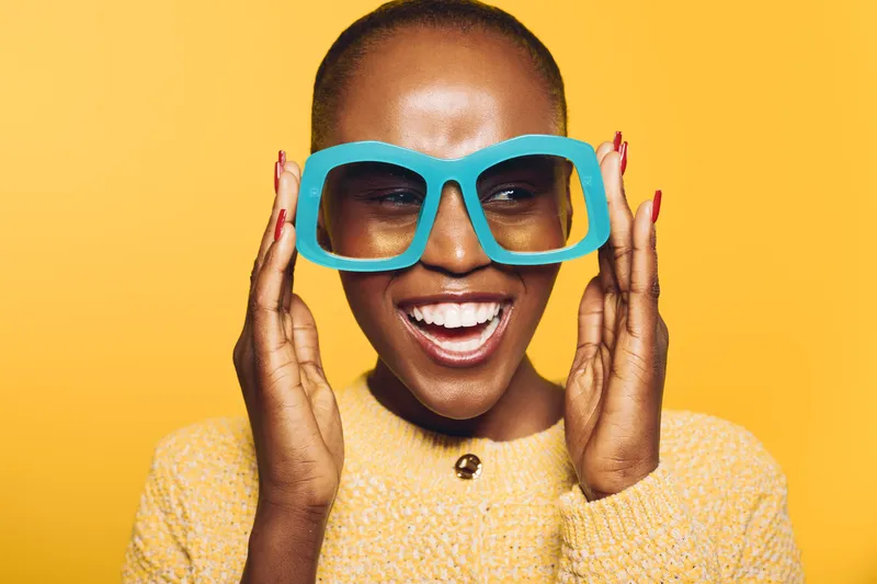 Happy smiling African woman with dark skin wearing oversized blue sunglasses and yellow sweater. Bright yellow background creates cheerful and vibrant mood. Concept of joy and fashion style.