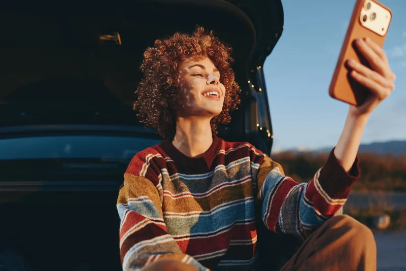 Woman lifestyle selfie from inside car, curly hair and striped sweater, smiling at golden sunset outdoors, relaxed posture, warm breeze atmosphere, scenic water horizon, evening light