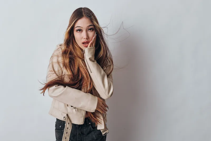 Young woman with long hair wearing light jacket and black pants posing against grey background with hand on face and thoughtful expression in studio portrait photography session.
